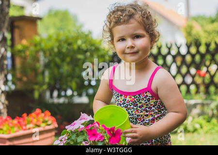 Süßes kleines Mädchen mit lockigem Haar lächelt, während die Bewässerung mit bunten Blumen im Garten Stockfoto