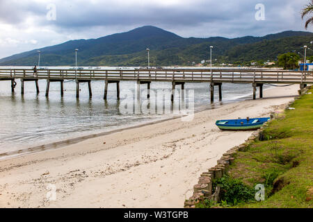 Detail der hölzernen Pier von Morrinhos Strand, mit hölzernen Angeln Boot und Strand Praia dos Zimbros im Hintergrund, Bombinhas, Santa Catarina Stockfoto