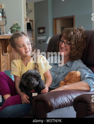 Eine lächelnde Frau hält ein fünf Jahre altes Mädchen in ihrem Schoß während der lächelnde Kind ihre Arme um ein Hund hat. Unposed Bild. USA Stockfoto
