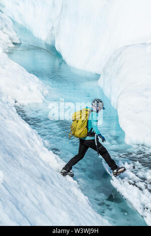 Weibliche Eiskletterer Schritt über eine tiefe Gletscherspalte mit klarem, blauem Wasser aus dem schmelzenden Eis der Matanuska Gletscher in Alaska gefüllt. Stockfoto