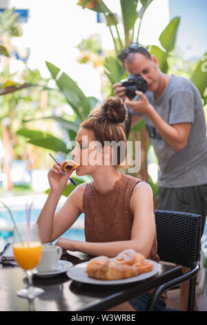Reizende junge Frau essen ein Brötchen Brot, während ihre Hausaufgaben - Bananenstauden und einen Pool im Hintergrund - ein Fotograf im Hintergrund Stockfoto