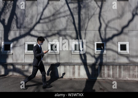 Ein junger japanischer Salaryman überprüft sein Smartphone, während er an einer Wand mit kleinen Fenstern und starken Baumschatten in Yoyogi Uehara, Tokio, Japan vorbeiläuft. Stockfoto