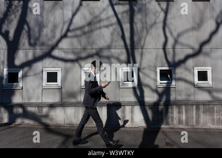 Ein junger japanischer Salaryman überprüft sein Smartphone, während er an einer Wand mit kleinen Fenstern und starken Baumschatten in Yoyogi Uehara, Tokio, Japan vorbeiläuft. Stockfoto