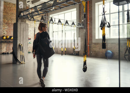 Morgen Workout. Rückansicht des jungen athletischen Frau, die ihr Sport Tasche und zur Seite schauen, während bei leeren Turnhalle stehen. Professionellen Sport. TRX Training. Training Stockfoto