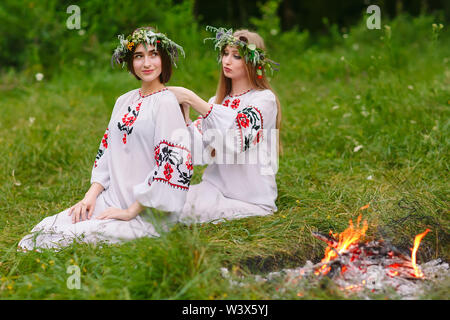 Hochsommer. Zwei Mädchen in der Slawischen Kleider weben Flechten der Haare in der Nähe des FIRE Stockfoto