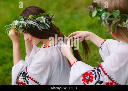 Hochsommer. Zwei Mädchen in der Slawischen Kleider weben Flechten der Haare in der Nähe des FIRE Stockfoto