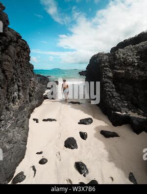 Fit männlich zu Fuß auf einem Sandstrand neben schwarzen Felsen Und erstaunliches Meer im Hintergrund Stockfoto