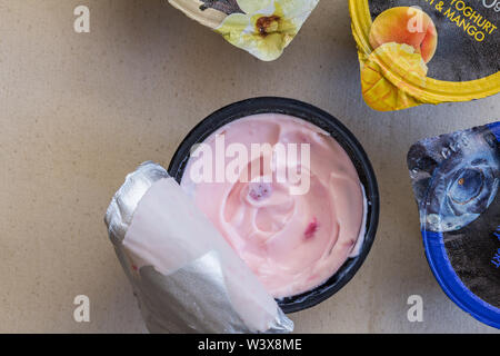 Erdbeer Joghurt in Kunststoff schwarz Cup auf Leinen Hintergrund mit Folie Deckel geöffnet und drei andere Früchte Joghurtbecher ungeöffnet - Ansicht von oben Hintergrund Stockfoto