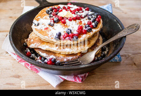 Hausgemachte Pfannkuchen gebraten auf schwarzem Gusseisen Skillet mit Beeren, Himbeeren, Preiselbeeren und Brombeeren auf hölzernen rustikalen braunen Hintergrund und Vin Stockfoto