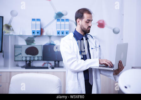 Junger Arzt in seinem Kabinett stehen und Arbeiten auf seinem Laptop. Arzt Kontrolle der Gesundheit von Patienten. Stockfoto