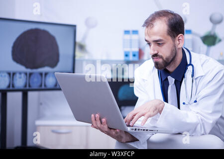 Attraktive bärtigen Doktor mit der Eingabe eines diagnosticin in der Patientendatei auf seinem Laptop. Attraktive Kardiologe Arbeiten am Laptop. Stockfoto