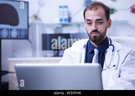Close up Portrait von gutaussehenden Arzt saß an seinem Schreibtisch und Arbeiten am Computer. Facharzt auf Gehirn Behandlung. Stockfoto