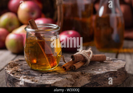 Glas Apfelwein mit Zimtstangen auf einem rustikalen Oberfläche mit roten Äpfeln im Hintergrund Stockfoto