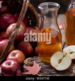 Karaffe von Apfelwein und Apple in die Hälfte geschnitten auf rustikalem Holz mit einem Korb rote Äpfel im Hintergrund verschüttet Stockfoto