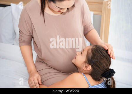 Süße Tochter Mutter zu umarmen und hören auf die Stimme des Babys im Bauch der Mutter prüfen und bei Mama. Schwangere und Eltern. Gesundheit und Med Stockfoto