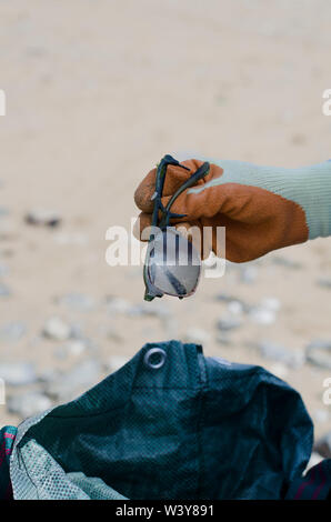 Hand mit Orange Handschuh Holding zerbrochene Gläser mit Plastiktüte im Hintergrund am Strand Stockfoto