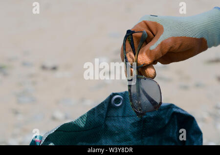 Hand mit Orange Handschuh Holding zerbrochene Gläser mit Plastiktüte im Hintergrund am Strand Stockfoto