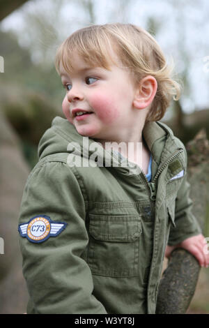 Little Boy (2/3 Jahre) trägt ein khaki Mantel, lächelnd und sah über seine Schulter: Mottisfont, Hampshire, UK. MODEL RELEASED Stockfoto