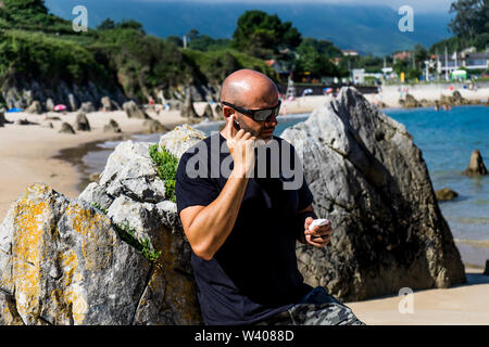 Glatzköpfige Mann am Strand mit Kopfhörern und Mobiltelefon. Stockfoto