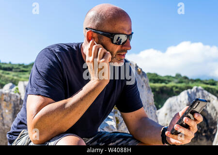 Glatzköpfige Mann am Strand mit Kopfhörern und Mobiltelefon. Stockfoto