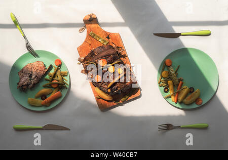 Tisch Einstellungen für Zwei, im Sonnenlicht und Schatten. Schweinesteak und gekochtem Gemüse auf grünen Platten. Oben Blick auf gesundes Mittagessen. Stockfoto