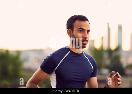 Portrait von aktiven tausendjährigen Mann Joggen in der Dämmerung mit einem urbanen Stadtlandschaft und den Sonnenuntergang im Hintergrund Stockfoto