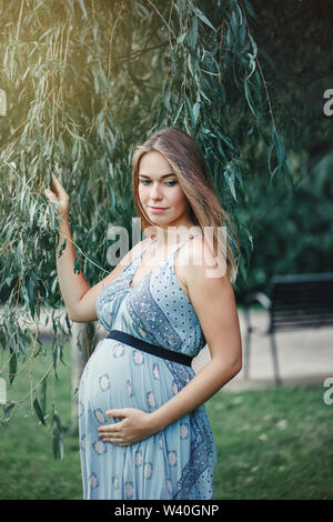 Glückliche gesunde Schwangerschaft und Mutterschaft. Portrait von Schwangeren jungen kaukasischen Frau mit langen blauen Kleid in Park außerhalb posieren. Schöne nachdenkliche Fe Stockfoto