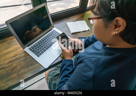 Asiatische Frau mit Handy während der Arbeit am Laptop Stockfoto