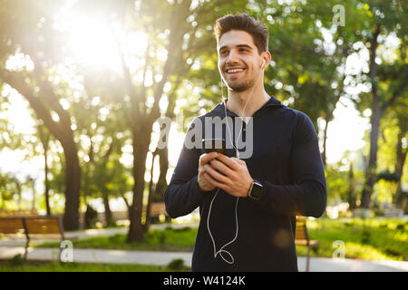 Bild von einer heiteren optimistischen jungen Sport Fitness Mann in Green Park Natur über das Handy Chatten Musik hören. Stockfoto