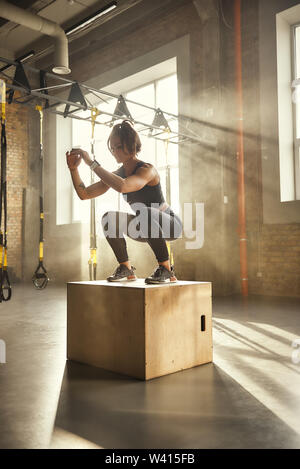 Mich herausfordernd. Seitenansicht der athletische Frau in Sportkleidung tun Hocke beim Stehen auf hölzernen Kasten im Fitnessstudio. Professionellen Sport. TRX Training. Training Stockfoto