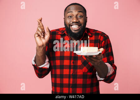 Portrait Of Happy aufgeregt afrikanischen Mann Shirt stehen isoliert, Halteplatte mit Geburtstagstorte, halten die Finger gekreuzt Stockfoto