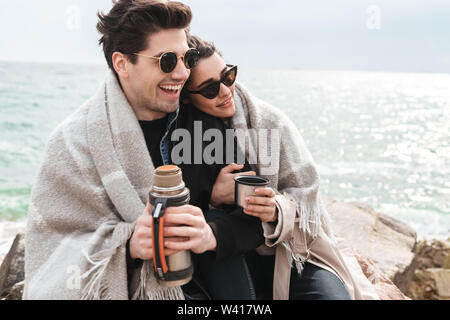 Glückliches junges Paar tragen Herbst Mäntel Zeit am Meer, sitzen in Decke zugedeckt, trinken Kaffee aus der Thermoskanne Stockfoto