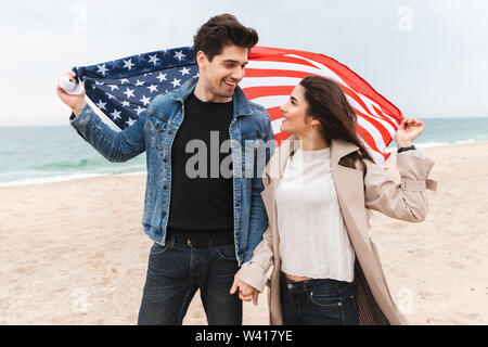 Happy reizende junge Paar Mäntel tragen, Wandern am Strand, Hand in Hand, die amerikanische Flagge Stockfoto