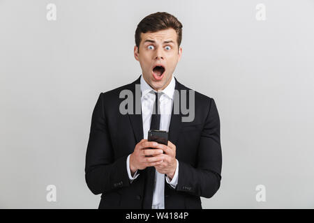 Bild von schockiert Yong Geschäftsmann in Anzug Tippen auf Handy und Kamera mit offenen Mund über grauer Hintergrund Stockfoto