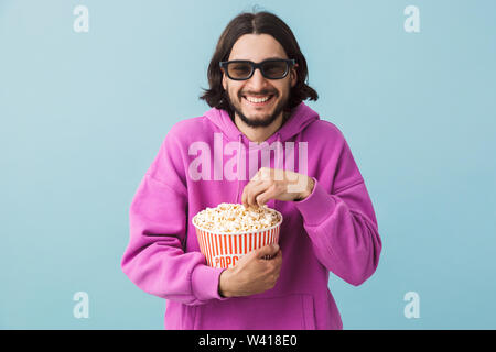 Portrait einer aufgeregt junge Bartgeier brünett Mann hoodie stehend über blauen Hintergrund isoliert, essen Popcorn, watching Film in 3D-Brille Stockfoto