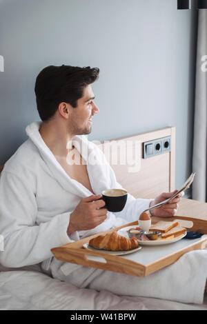 Foto junge lächelnde Mann mit weißer Bademantel frühstücken und Zeitung lesen, beim Sitzen auf dem Bett im Hotel Apartment Stockfoto