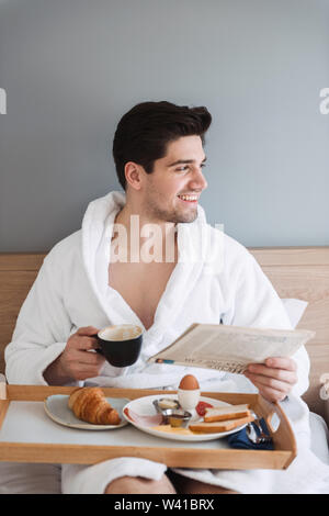 Foto der Jungen glücklich Mann mit weißer Bademantel frühstücken und Zeitung lesen, beim Sitzen auf dem Bett im Hotel Apartment Stockfoto