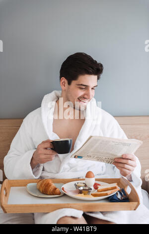 Foto der jungen attraktiven Mann mit weißer Bademantel frühstücken und Zeitung lesen, beim Sitzen auf dem Bett im Hotel Apartment Stockfoto