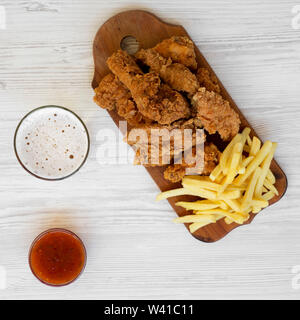 Lecker fast food: Fried Chicken Drumsticks, würzige Flügel, Pommes frites und Chicken Fingers mit Sauer-süßen Soße und Glas kaltes Bier auf einem weißen Woode Stockfoto