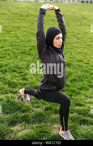 Portrait von muslimischen fitness Frau in religiösen Schwarz hijab ihren Körper stretching beim Workout im Green Park im Freien gekleidet Stockfoto