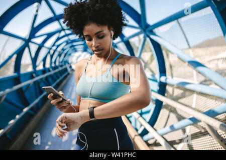 Sportlerin, die auf Smart Watch schaut und ihr Smartphone in der anderen Hand hält, draußen. Fitness-Frau richtet ihre Smart Watch für ihren Lauf ein. Stockfoto