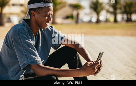 Afrikanische amerikanische jungen Kerl sitzt im Freien und mit einem Mobiltelefon. Junger Mann eine sms mit seinem Handy und lächelnd, Stockfoto