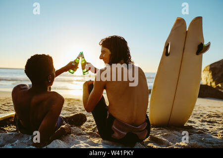 Ansicht der Rückseite zwei junge Surfer toasten Bier am Strand. Junge Männer feiern am Strand mit Surfbrett auf der Seite. Stockfoto