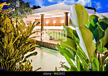 Gastgarten oder Terrasse Blick durch die Blätter der ausgefallenen Pflanzen über den Pool. Der Ort ist mit Lichtern in der Nacht beleuchtet Stockfoto