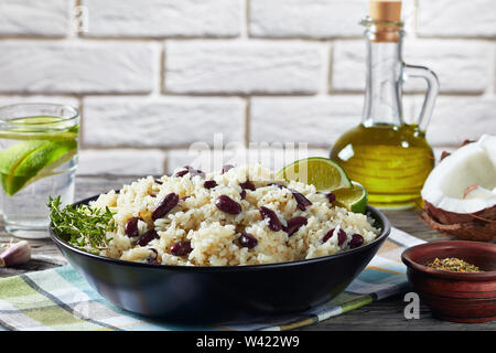 Karibik, Reis und rote Bohnen mit Kokosmilch, gewürzt mit Knoblauch, Zwiebeln und kreolischen Spice in einem schwarzen Schüssel mit Zutaten und ein Glas wa gekocht Stockfoto