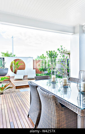 Terrasse im Freien mit Rattan Stühle auf dem Holzboden neben der Glasscheibe Tabelle, Es gibt Kerzen neben dem Garten Boden mit Steinen, es Stockfoto