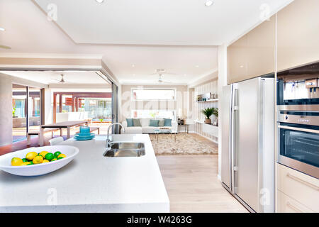 Der Innenraum des Luxus haus mit Geschirr, weiß Zähler nach oben Tabelle mit Obst auf einem weißen Teller in der Nähe von einem silbernen Waschbecken und Wasserhahn, ein Wohnzimmer ro Stockfoto