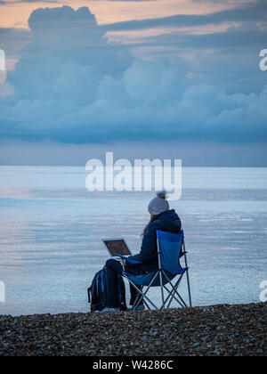 Eine junge Frau sitzt auf einem Stuhl an einem peeble beach Remotes Arbeiten mit Ihrem Laptop Stockfoto
