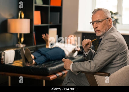 Stattliche schwere grauhaarige Psychiater sitzt in seinem Büro Stockfoto