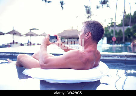 Junger Mann relaxen am Pool, mit Handy. Arbeit und Entspannung Stockfoto